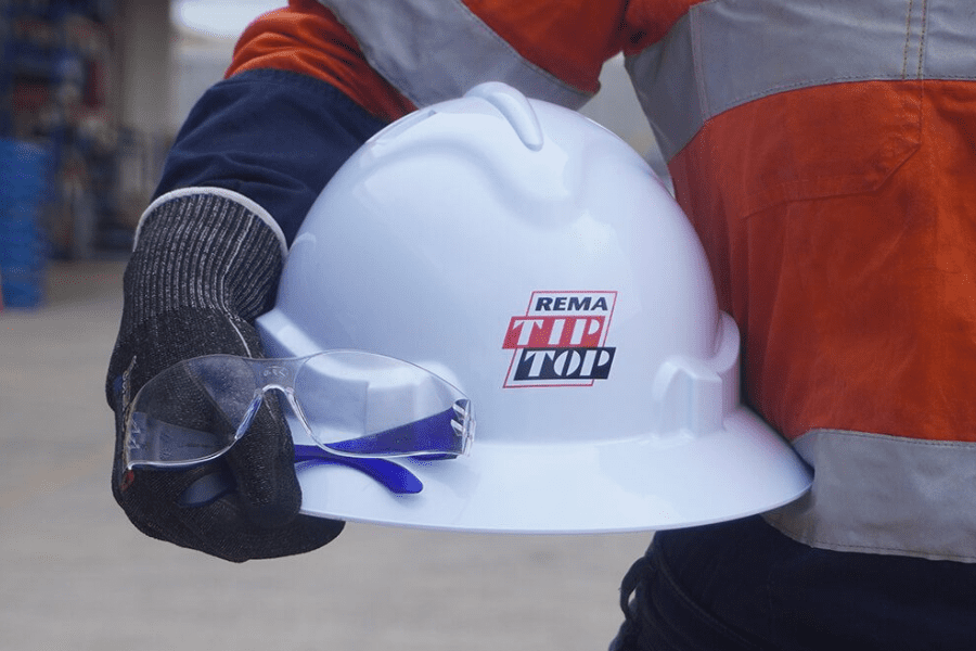 A worker in an orange hi-vis jacket holds a white safety helmet with goggles and a "rema tip top" logo in one hand, symbolizing career development.