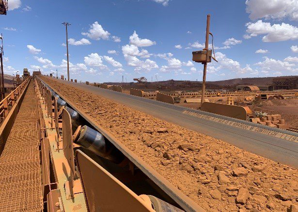 A conveyor belt transports loose soil in an industrial mining facility under a partly cloudy sky.