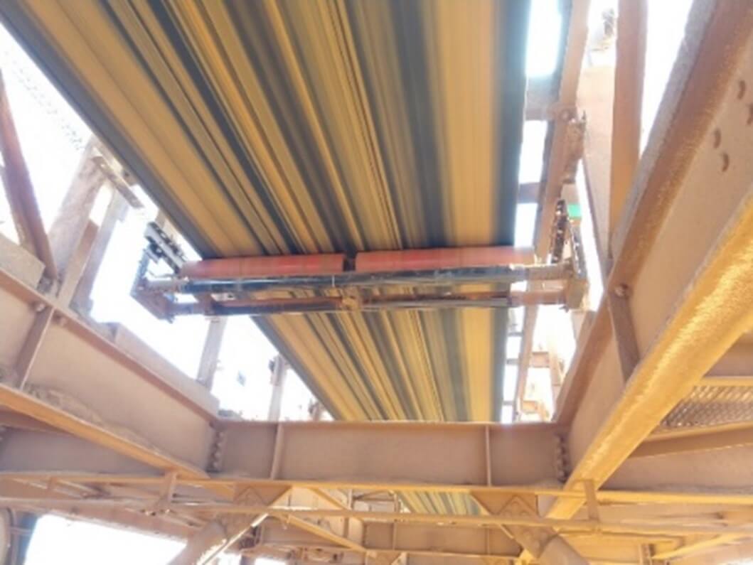 View from below a conveyor belt system, showing the underside of a moving belt with structural support beams and mechanical components visible.