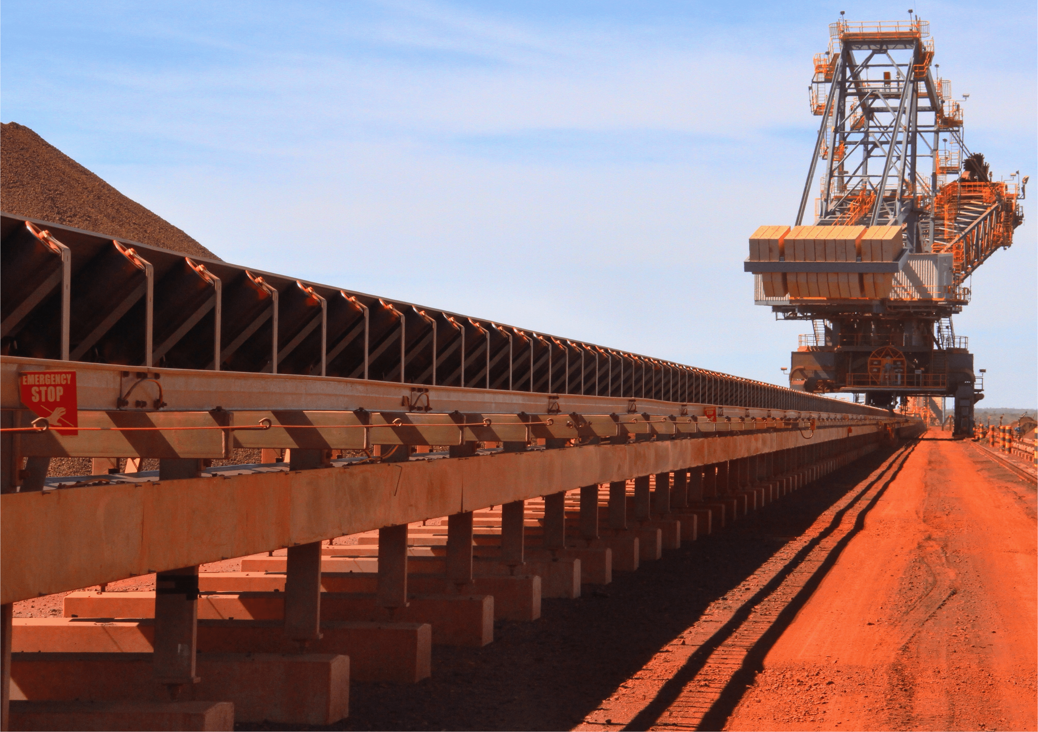 A large conveyor system transports materials in an open-pit mining site, with a sign on the left indicating an emergency stop mechanism. The ground and equipment are covered in reddish-brown dust.