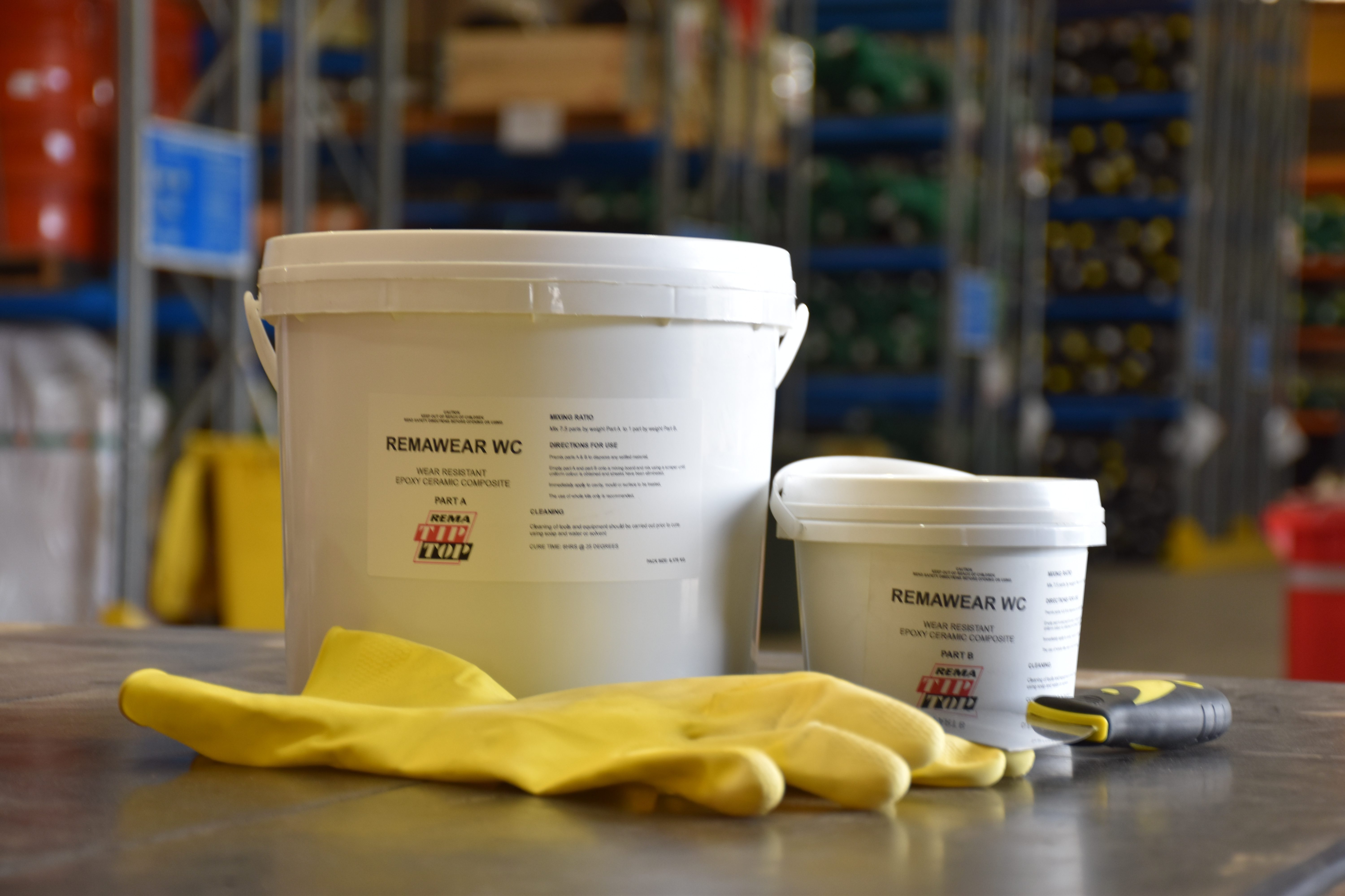 Two containers labeled "REMAWEAR WC" are placed on a table, along with a pair of yellow gloves and a tool. The background is a warehouse with shelves.