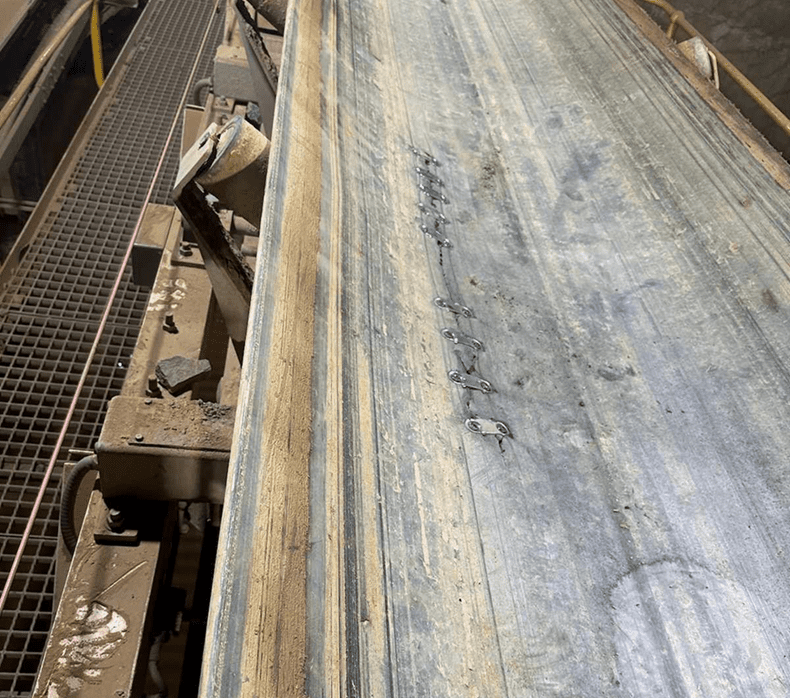 A worn industrial conveyor belt with visible patchwork and repair stitches running along its center. Metal grating and machinery parts are partially visible to the side.