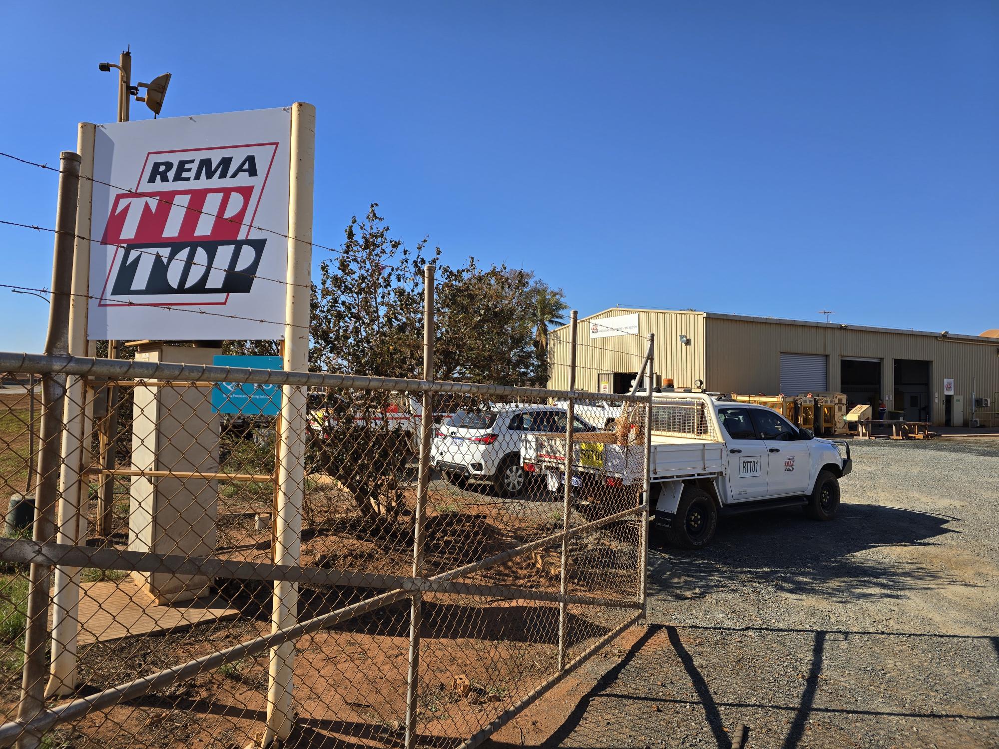 A fenced entrance with a "REMA TIP TOP" sign leads to a building, with parked vehicles visible inside a gravel yard area under a clear blue sky.