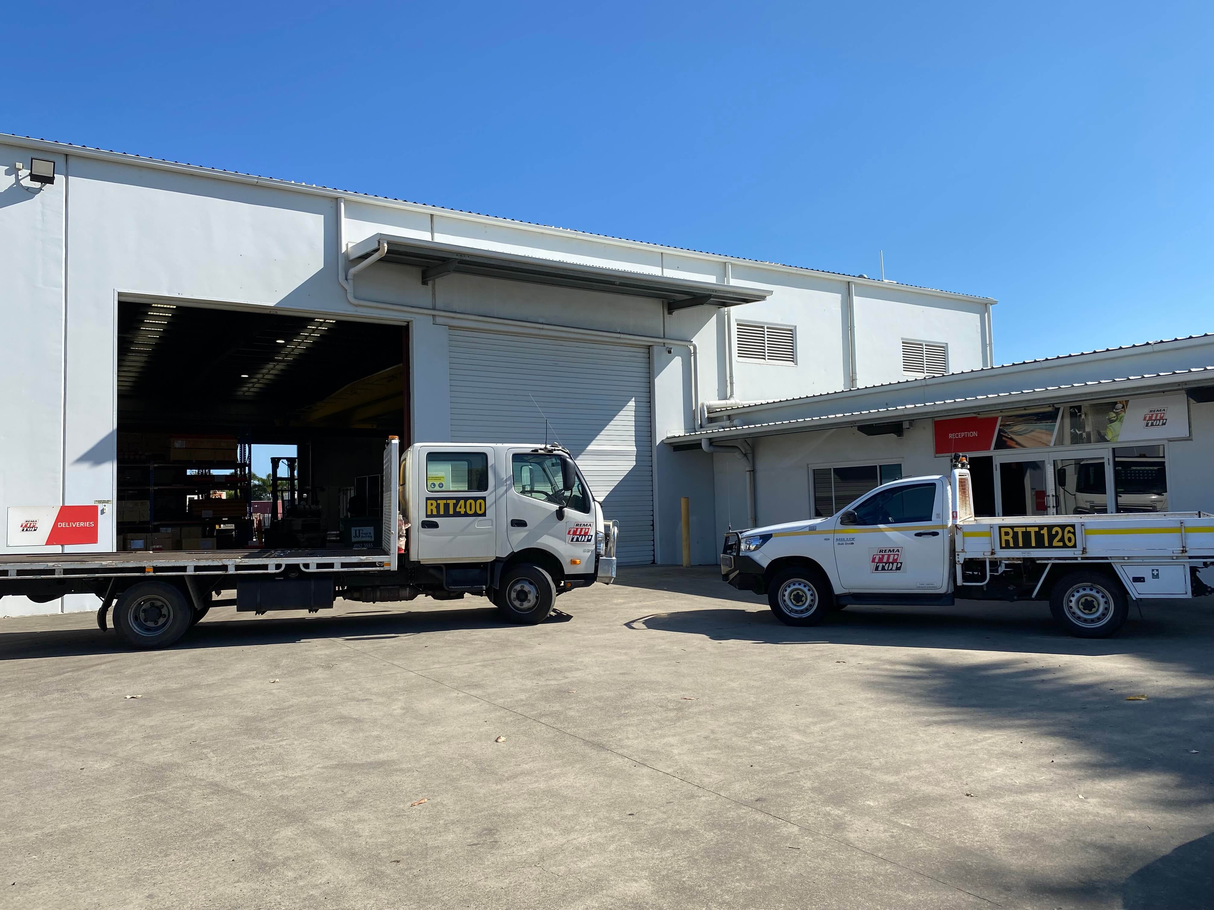 Two work trucks with the labels "RTT400" and "RTT126" are parked outside an industrial garage under a clear sky. The garage door is partially open, revealing some equipment inside.