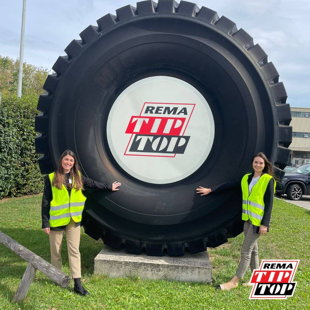 Two people in yellow safety vests stand beside a giant tire with the REMA TIP TOP logo, on a grassy area.