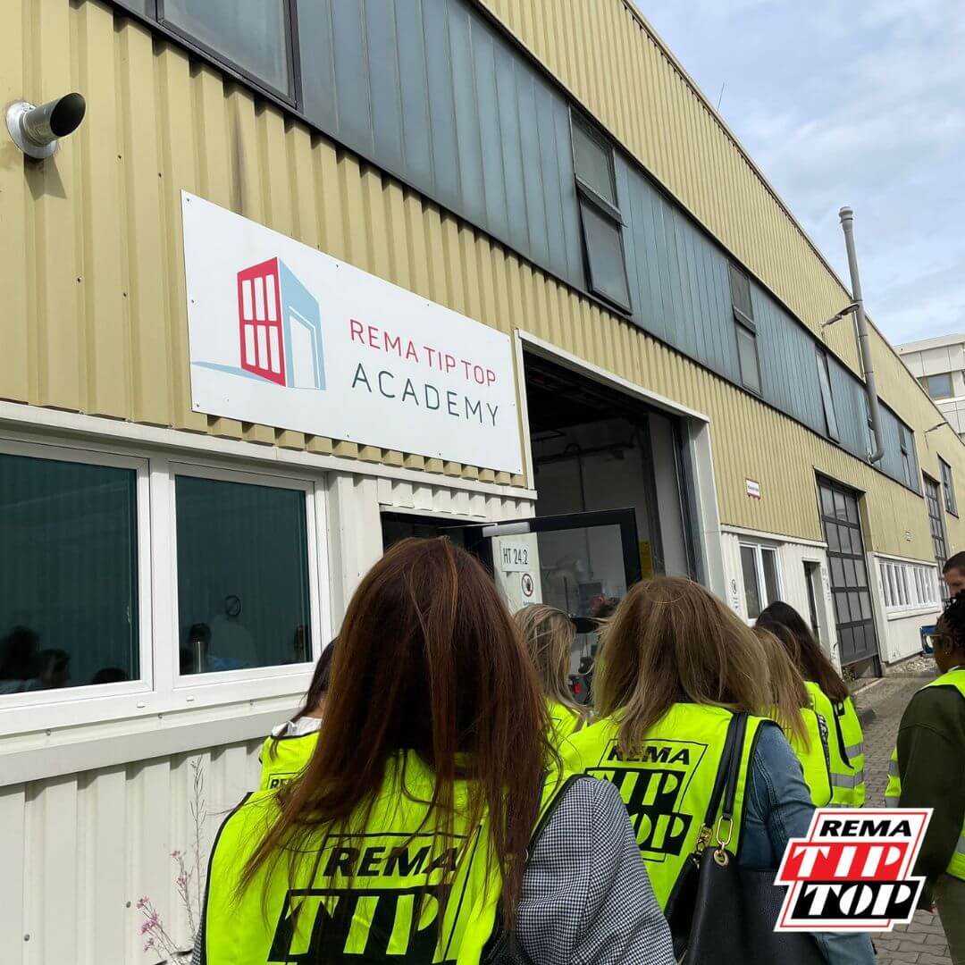 A group of people wearing safety vests enters a building labeled "REMA TIP TOP Academy.