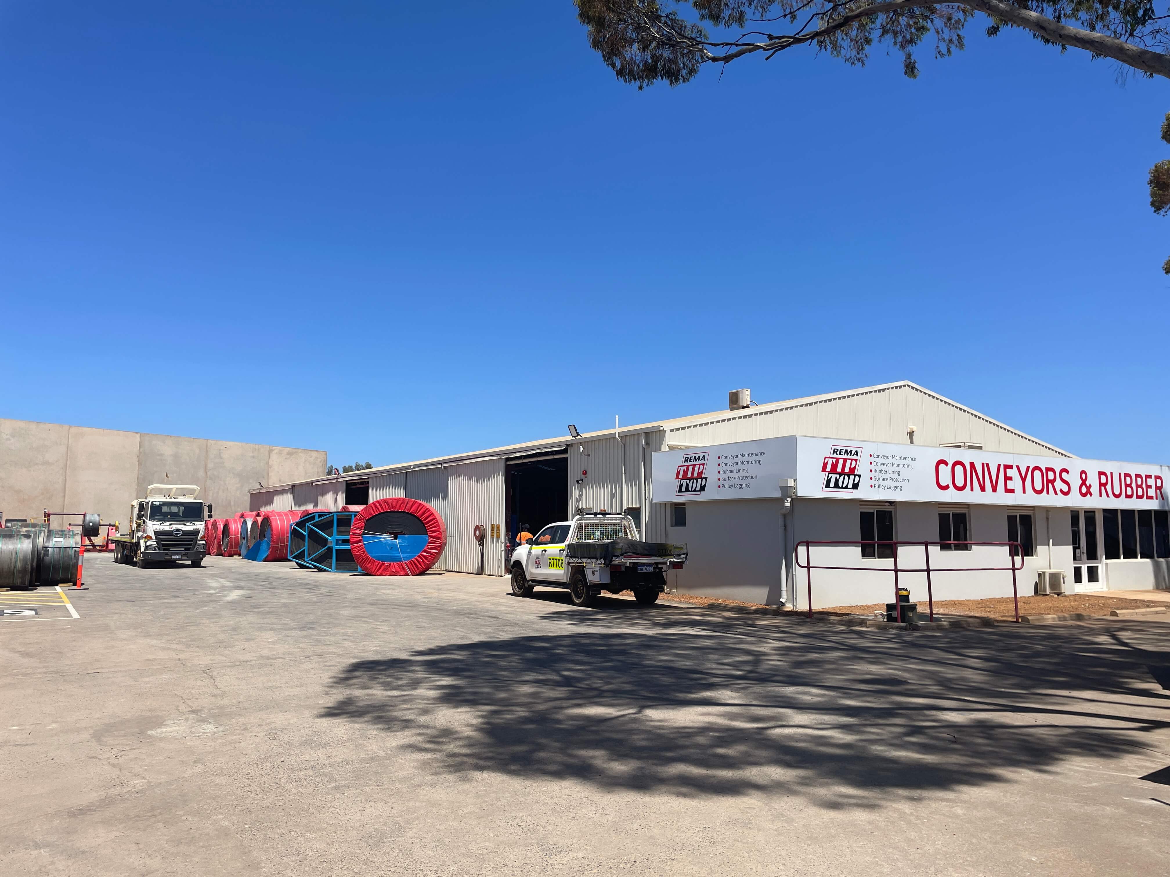 Industrial building with a sign reading "Conveyors & Rubber," trucks parked in front, and large coils of material visible outside.