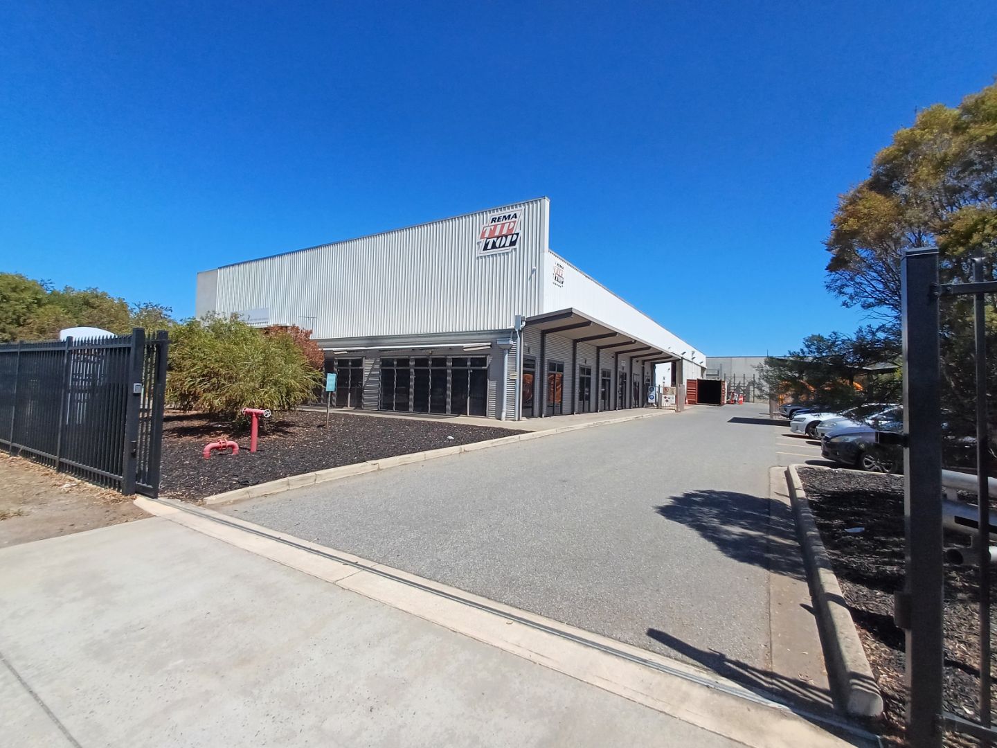 Commercial warehouse with a logo on the upper front, surrounded by a fence. Trees and parked cars are visible on the side, with a clear blue sky overhead.