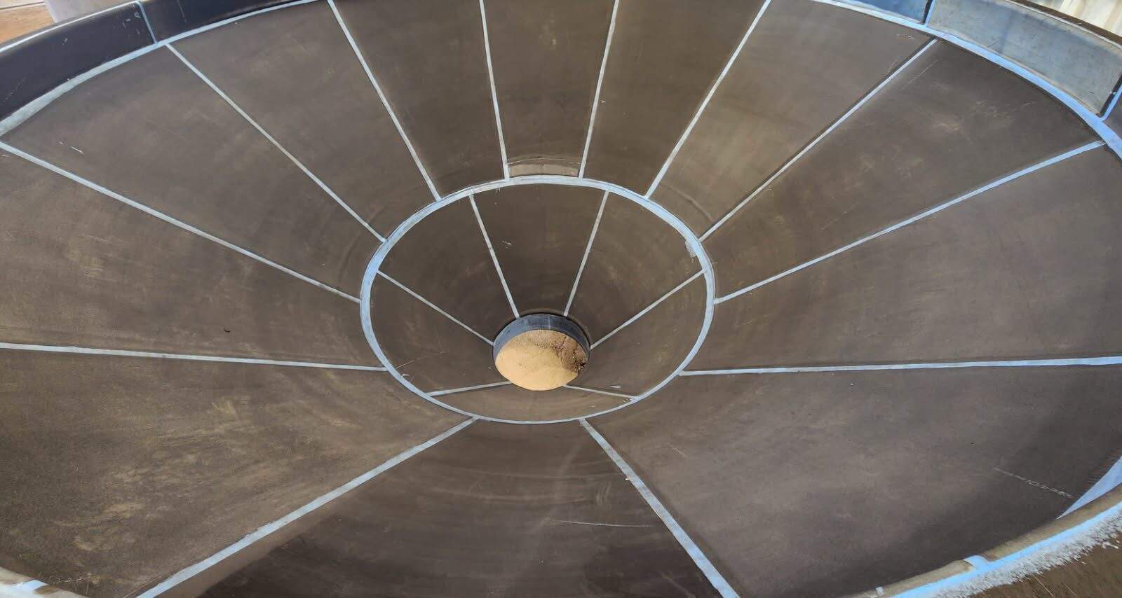 Large metal funnel structure with radial partitions, viewed from above, converging at a central circular opening with a beige object inside.