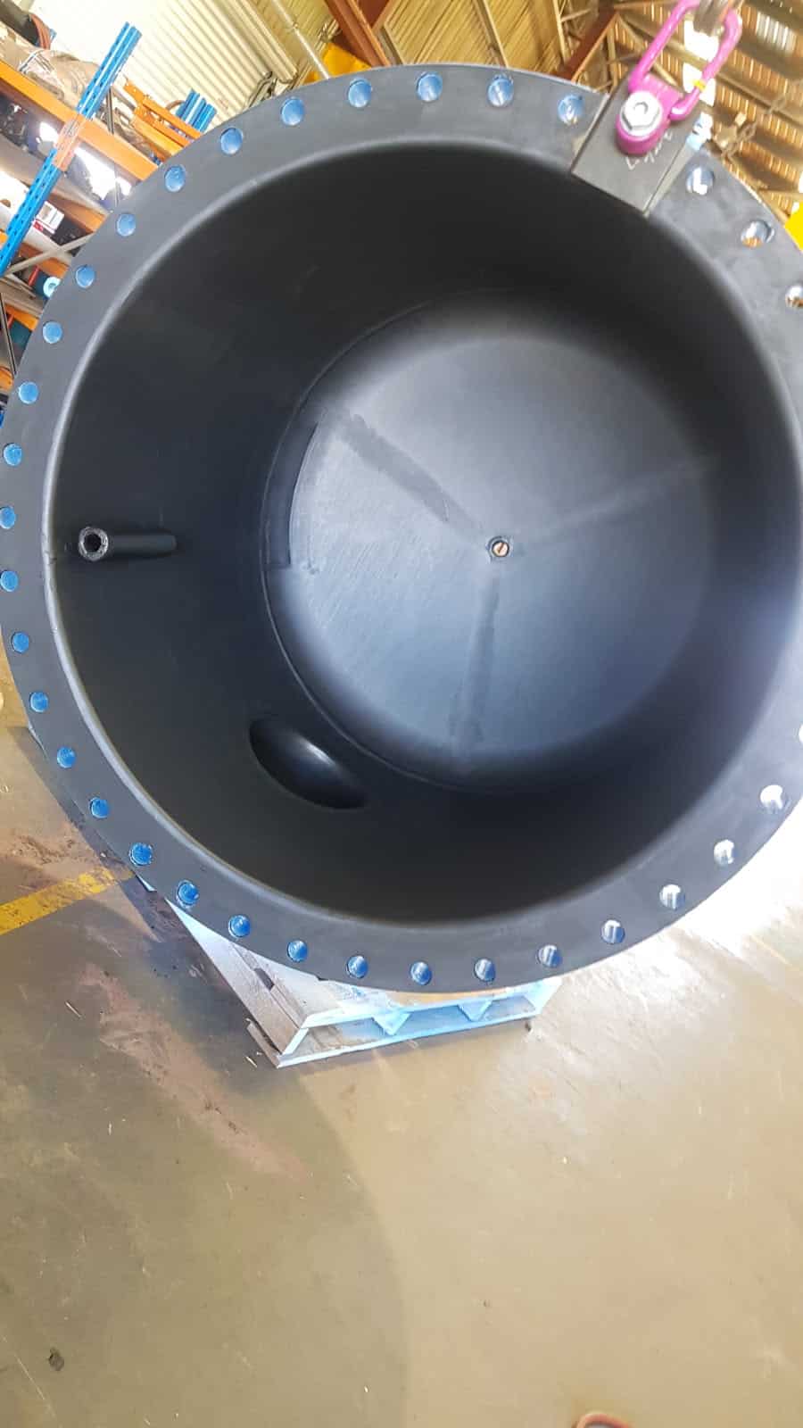 Large, empty black industrial tank with bolts around the rim, an inlet pipe on one side, and a pallet underneath, situated in a warehouse setting.