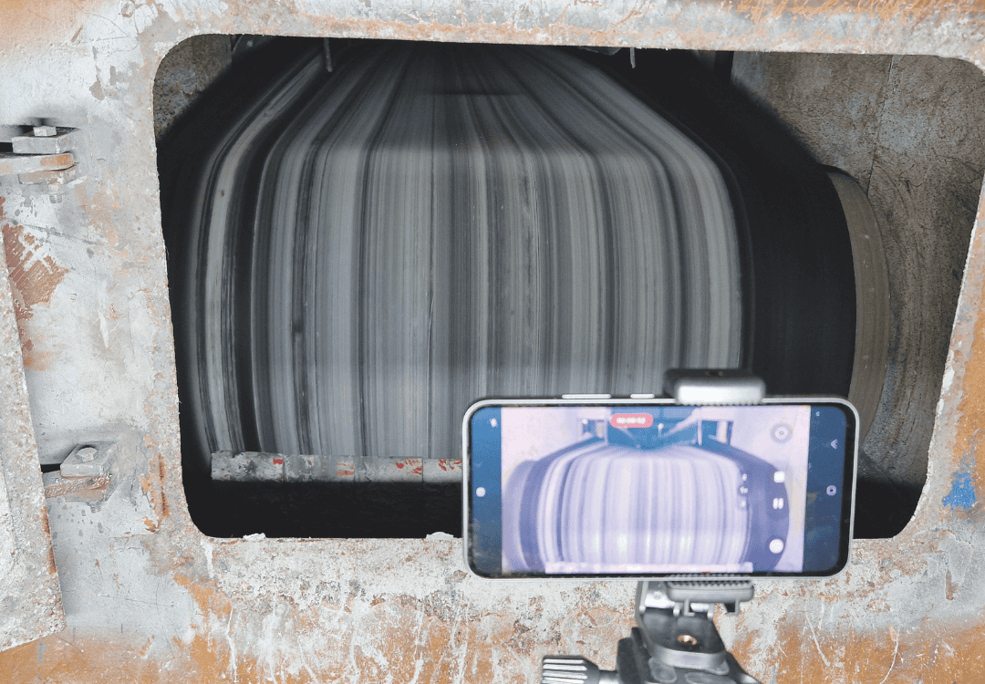 A smartphone on a tripod records a large industrial drum with black and white streaks inside a metal enclosure.