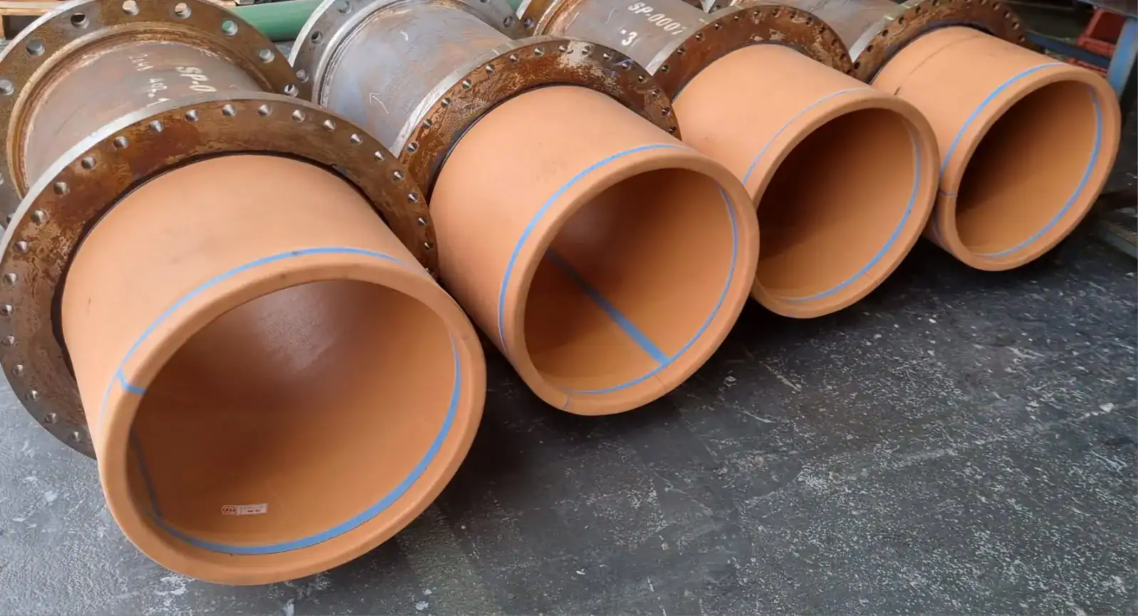 Four large, brown industrial pipes with metal flanges are arranged side by side on a dark, textured floor. The pipes have a blue stripe along their inner surface and appear clean and unused.
