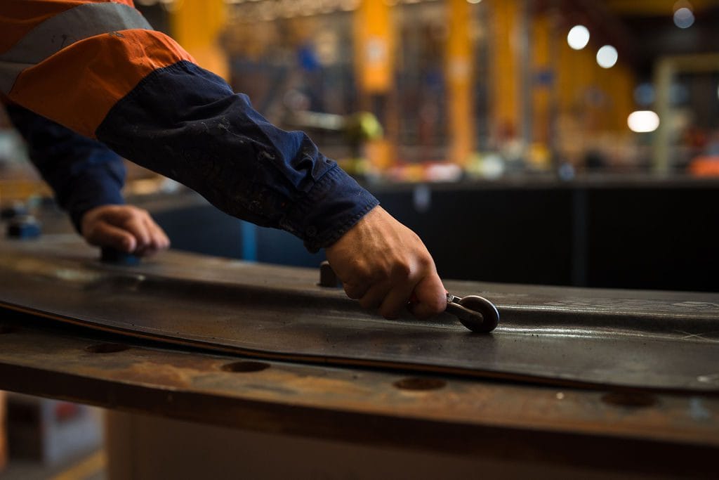 A person wearing a high-visibility safety jacket uses a measuring tool on a metal surface with rubber lining in an industrial workshop.