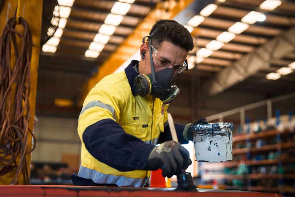 A worker in protective gear paints a metal surface with black paint inside an industrial facility, preparing it for rubber lining. Shelves with equipment are visible in the background.