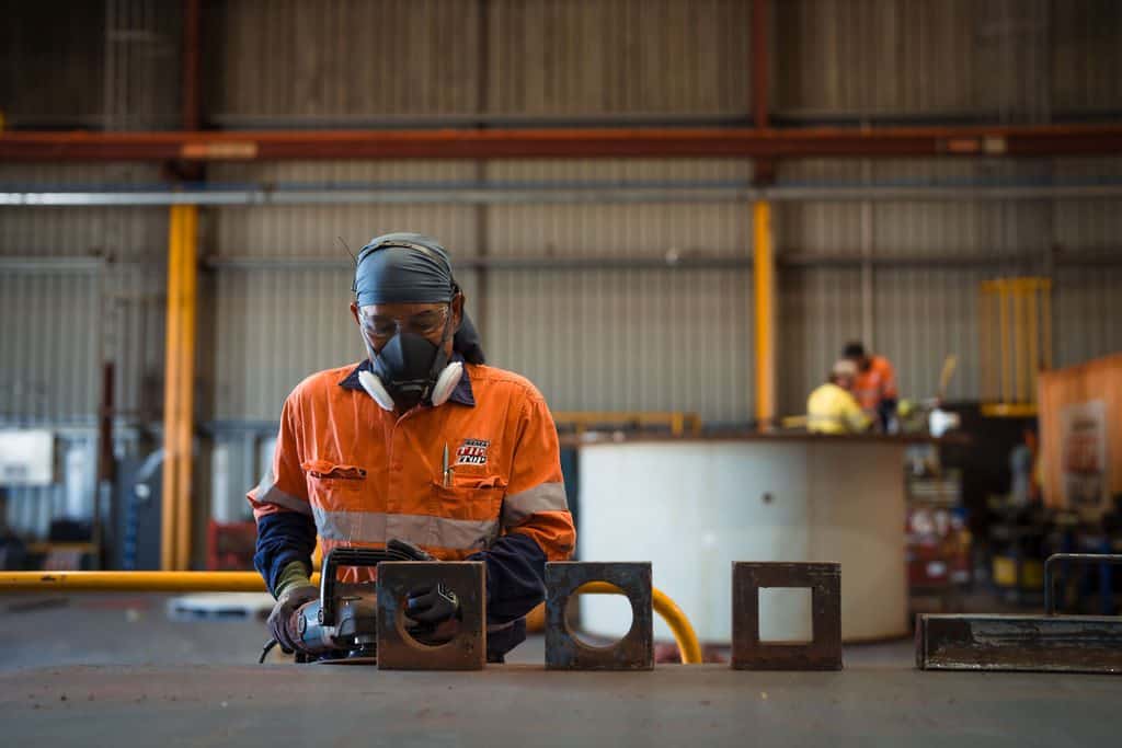 A worker wearing safety gear operates machinery in an industrial workshop, where metal plates and equipment with rubber lining are visible on the table and in the background.